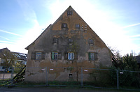 Der massive Westgiebel ist zwar als Bauteil noch vollständig überliefert, er steht jedoch auf Grund des eingestürzten Daches gegenwärtig ohne Windsicherung da. Die Westseite / Außenseite der Wand ist in einem Zuge aufgemauert und zeigt keine Versätze, Vor oder Rücksprünge oder Anzeichen von Gliederungselementen. Zusätzlich zum Wetterschutz gegen Westen ist denkbar, dass die hinter der massiven Erdgeschosswand vermuteten Lagerräume im Erdgeschoss auf diese Weise kühl gehalten werden konnten, da das Gebäude auf Grund des felsigen Untergrundes laut Aussage der Eigentümerin keinen Keller besitzt. / Haus Osiander in 74549 Wolpertshausen-Rudelsdorf (05.04.2024 - Schäfer, Ferdinand M. (SCHÄFER.PARTNER PartG Architur.Städtebau.Denkmalpflege, Schwäbisch Hall))